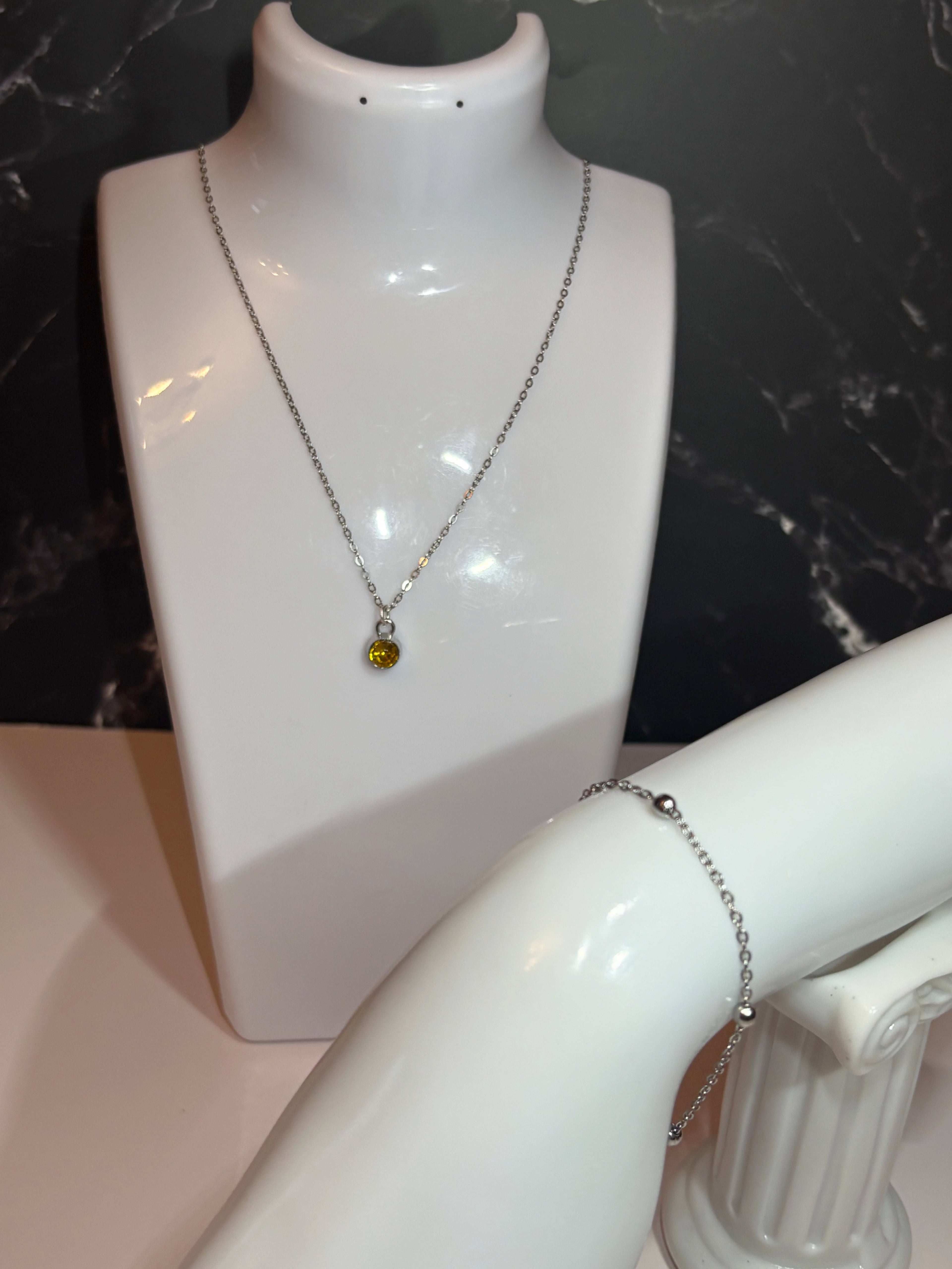 A silver-toned chain necklace with a small round November birthstone pendant displayed on a white jewelry bust, alongside a silver ball chain bracelet on a white arm form against a dark marble-patterned background