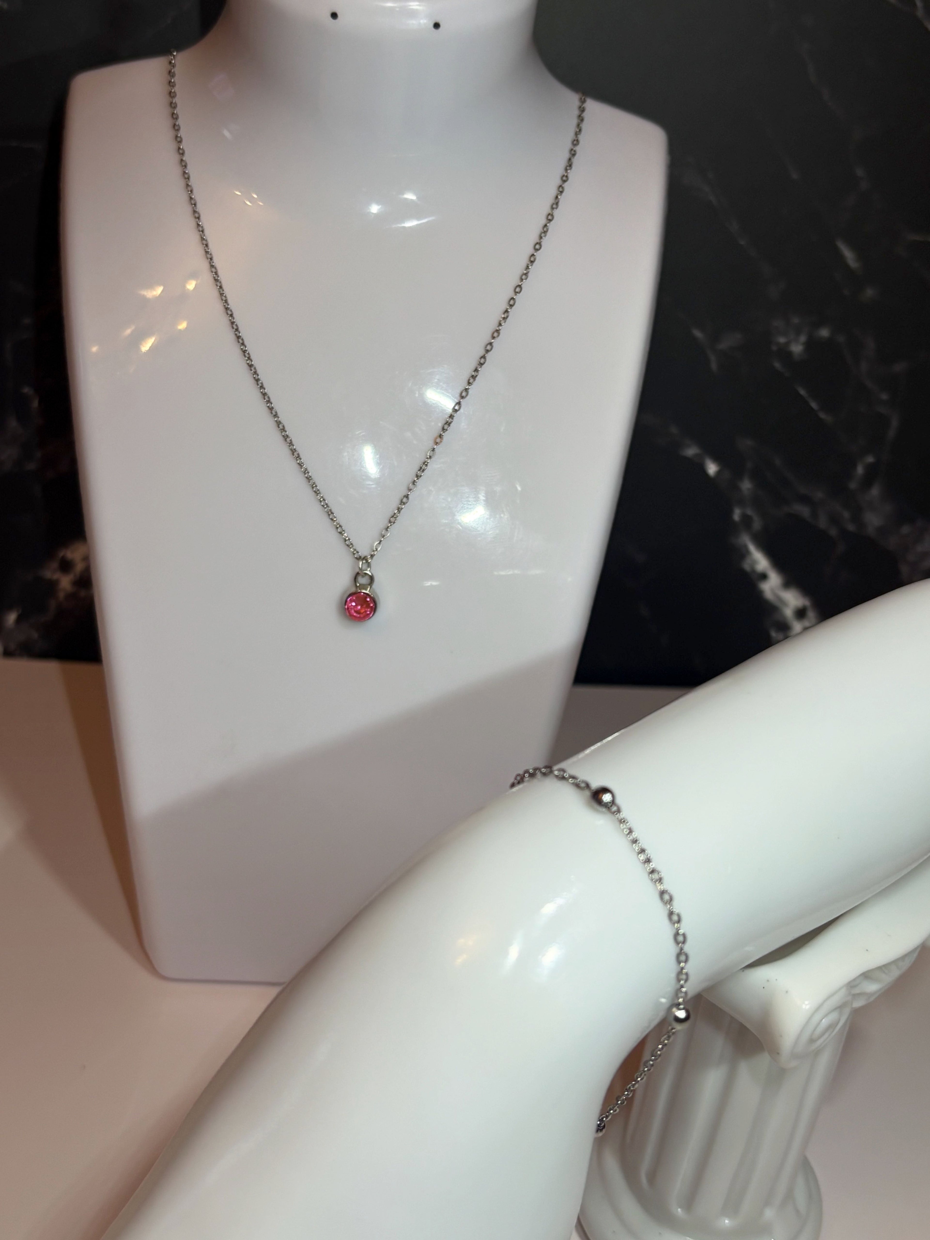 A silver chain necklace with a small pink birthstone pendant displayed on a white mannequin bust, with a silver ball chain bracelet shown on a mannequin arm against a dark marble background.