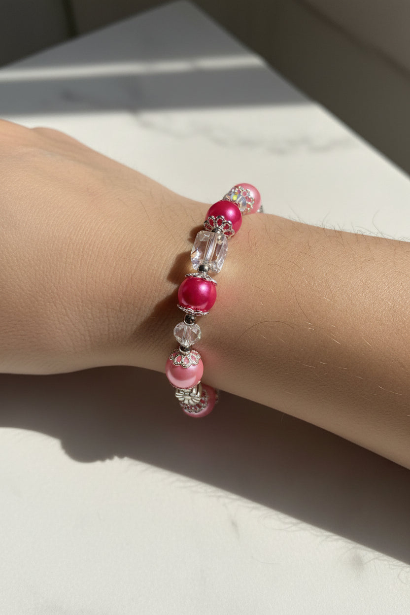 A beaded stretch bracelet featuring alternating pink glass pearls, bright magenta beads, clear faceted crystal beads, and silver-toned metal spacer details, displayed on a plain light beige background.