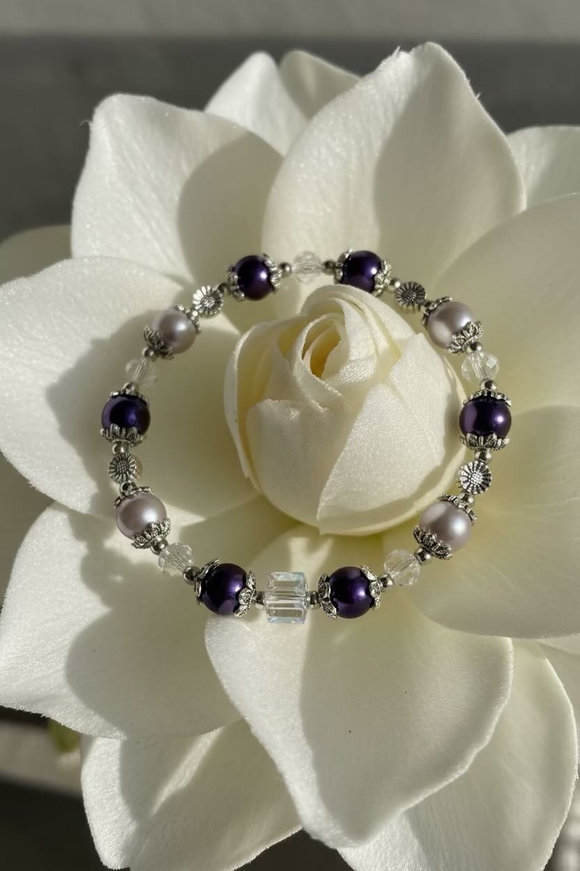 A beaded stretch bracelet with dark purple, silver, and lilac glass pearl beads, displayed on the center of a large white flower.