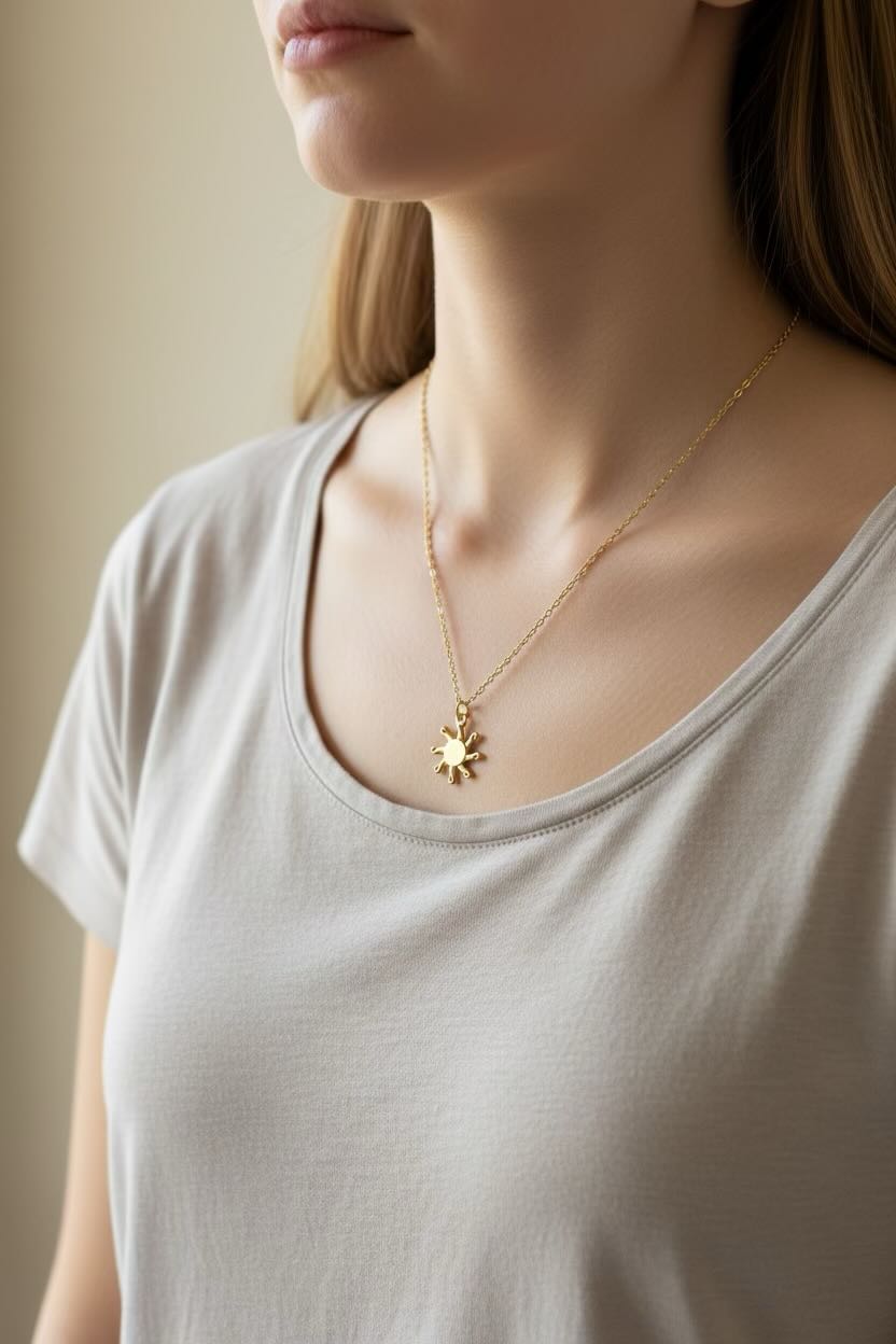 A close-up of a woman wearing a delicate gold chain necklace with a small radiant sun pendant, paired with a light beige scoop-neck t-shirt.