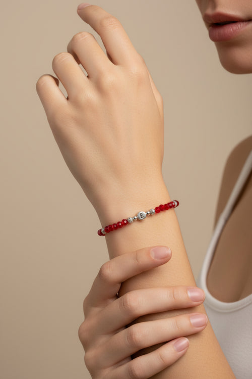 A beaded bracelet with red faceted beads, small silver spacer beads, and a central silver disc charm engraved with the letter 'E', displayed on a model