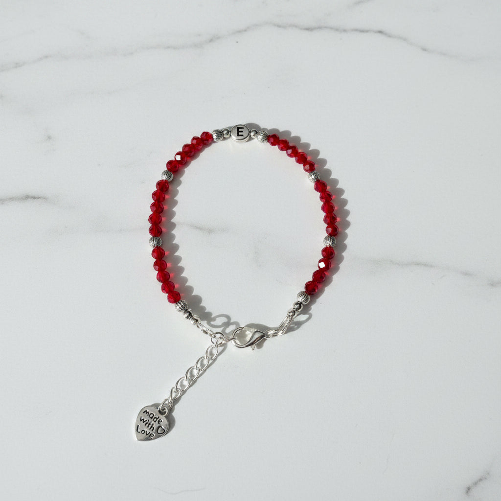 A beaded bracelet with red faceted beads, silver spacer beads, a silver lobster clasp, and a dangling heart charm engraved with letter 'E', displayed on a neutral gray background.