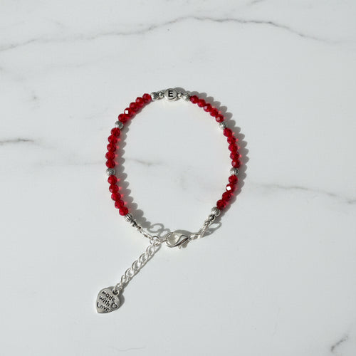 A beaded bracelet with red faceted beads, silver spacer beads, a silver lobster clasp, and a dangling heart charm engraved with letter 'E', displayed on a neutral gray background.