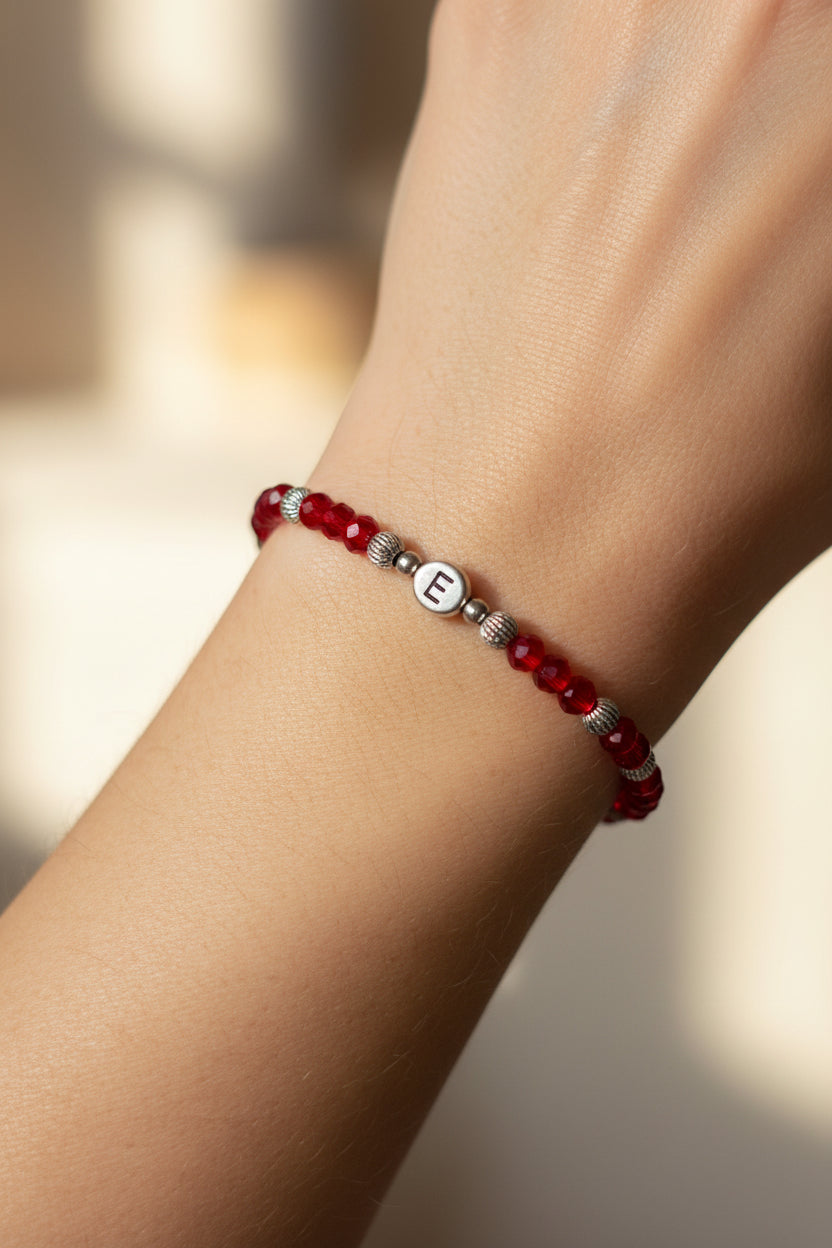 A red beaded bracelet with silver spacer beads and a central silver letter charm spelling 'E', displayed on a white model's arm