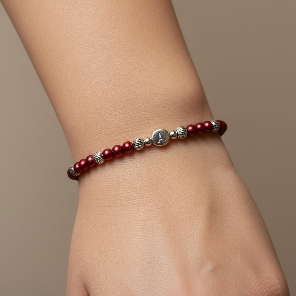 A delicate beaded bracelet with red pearls and silver spacer beads, featuring a central silver disc engraved with the letter 'A', displayed on a model
