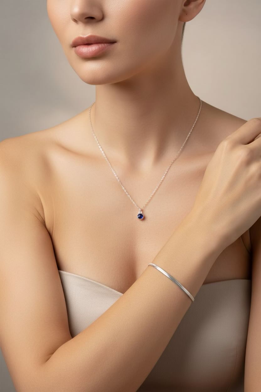 A close-up of a model wearing a delicate silver necklace with a small round blue birthstone pendant, paired with a silver flat snake chain bracelet, set against a soft beige background.