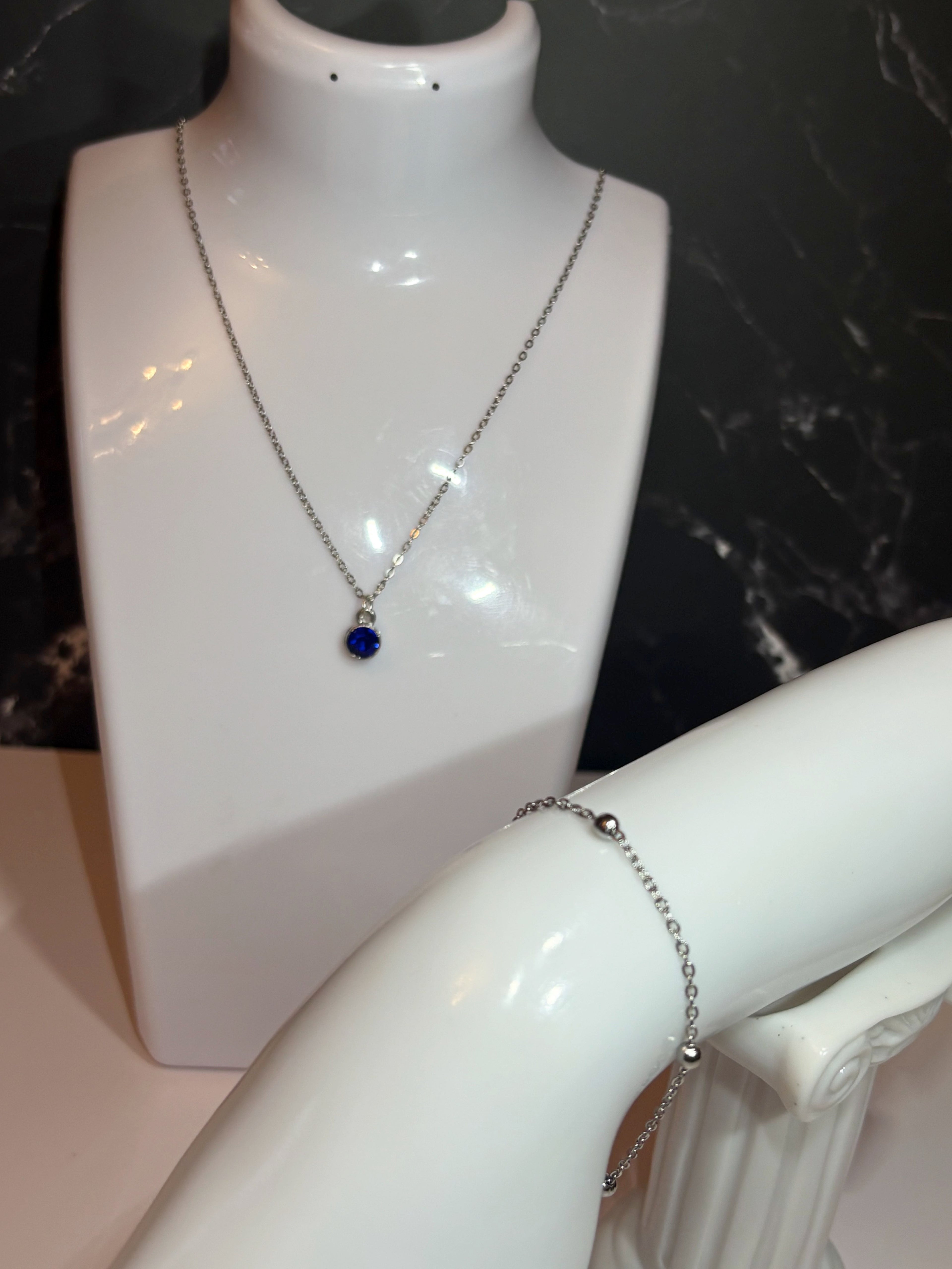 A silver chain necklace with a small blue birthstone circular pendant displayed on a white jewelry bust, with a silver ball chain bracelet shown on another white bust arm against a dark marble background.