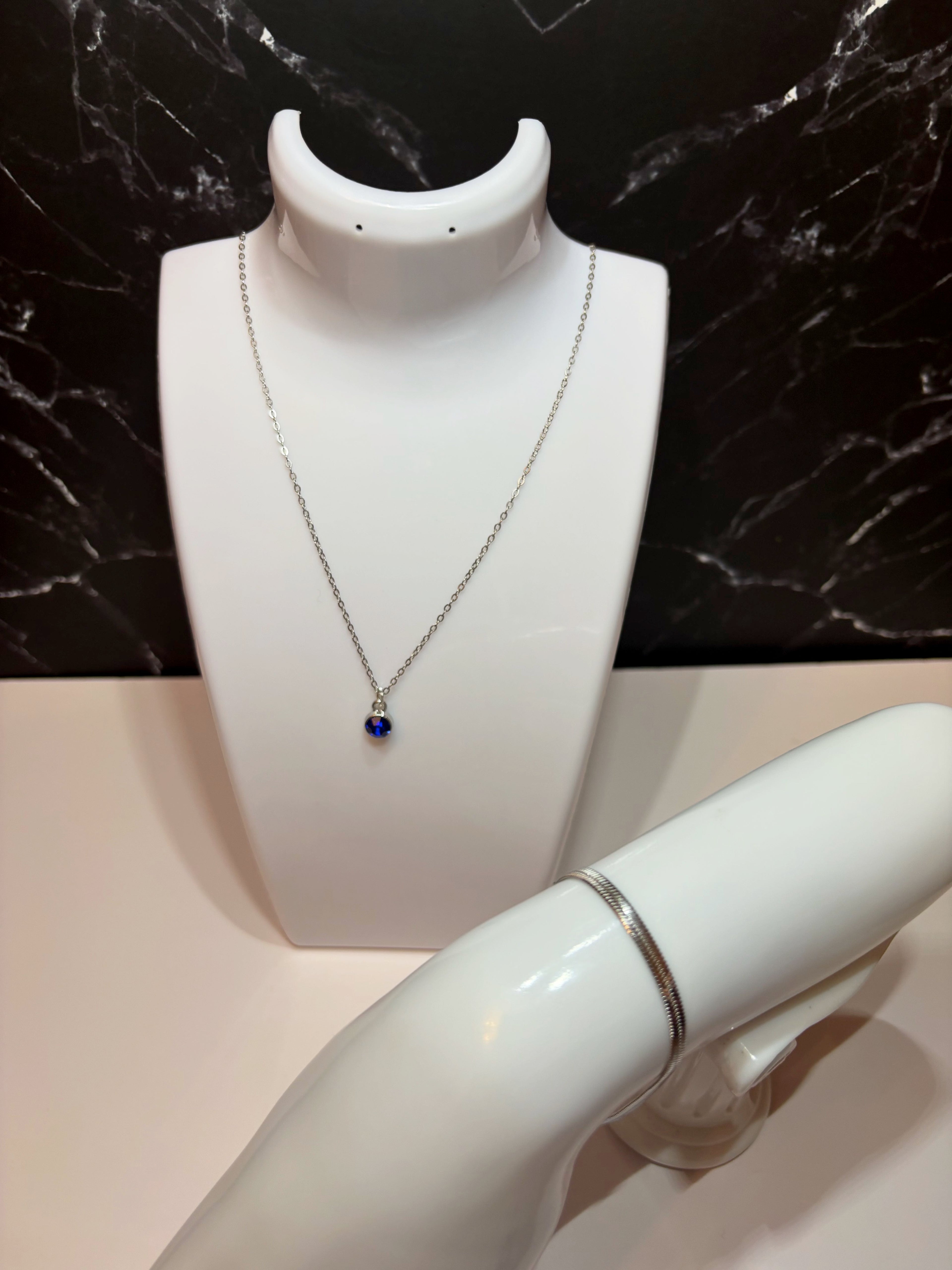 A silver chain necklace with a small blue sapphire birthstone pendant displayed on a white jewelry bust, with a silver flat snake chain bracelet also shown on a white arm form against a black marble background.