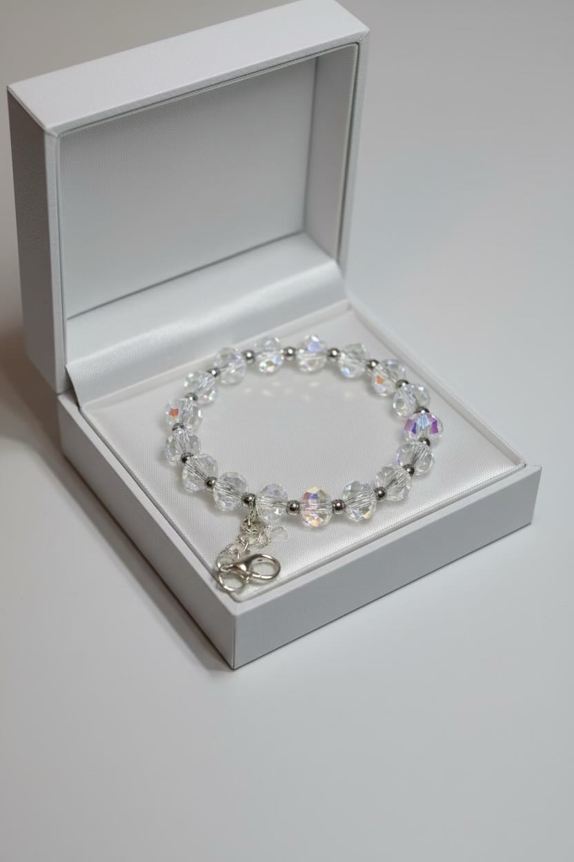 A beaded bracelet with clear faceted beads, iridescent accents, and a small silver charm, displayed inside an open white jewelry box on a light gray background.