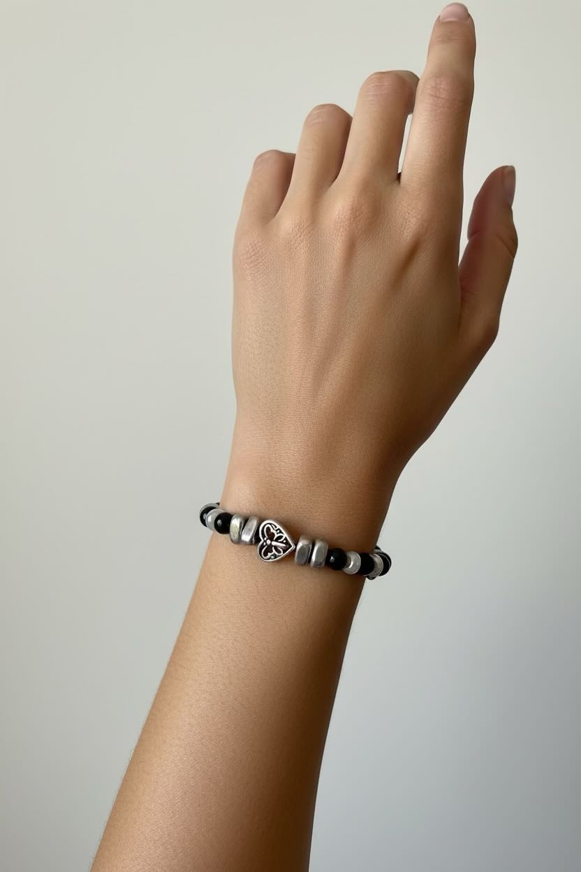 A close-up of a person's wrist wearing a beaded bracelet with a central silver-toned heart charm featuring a heart design, set against a plain light gray background.