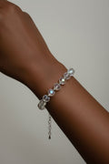 A close-up of a person's wrist wearing a delicate silver-toned bracelet adorned with faceted clear crystal beads and a small dangling chain, set against a plain light gray background.