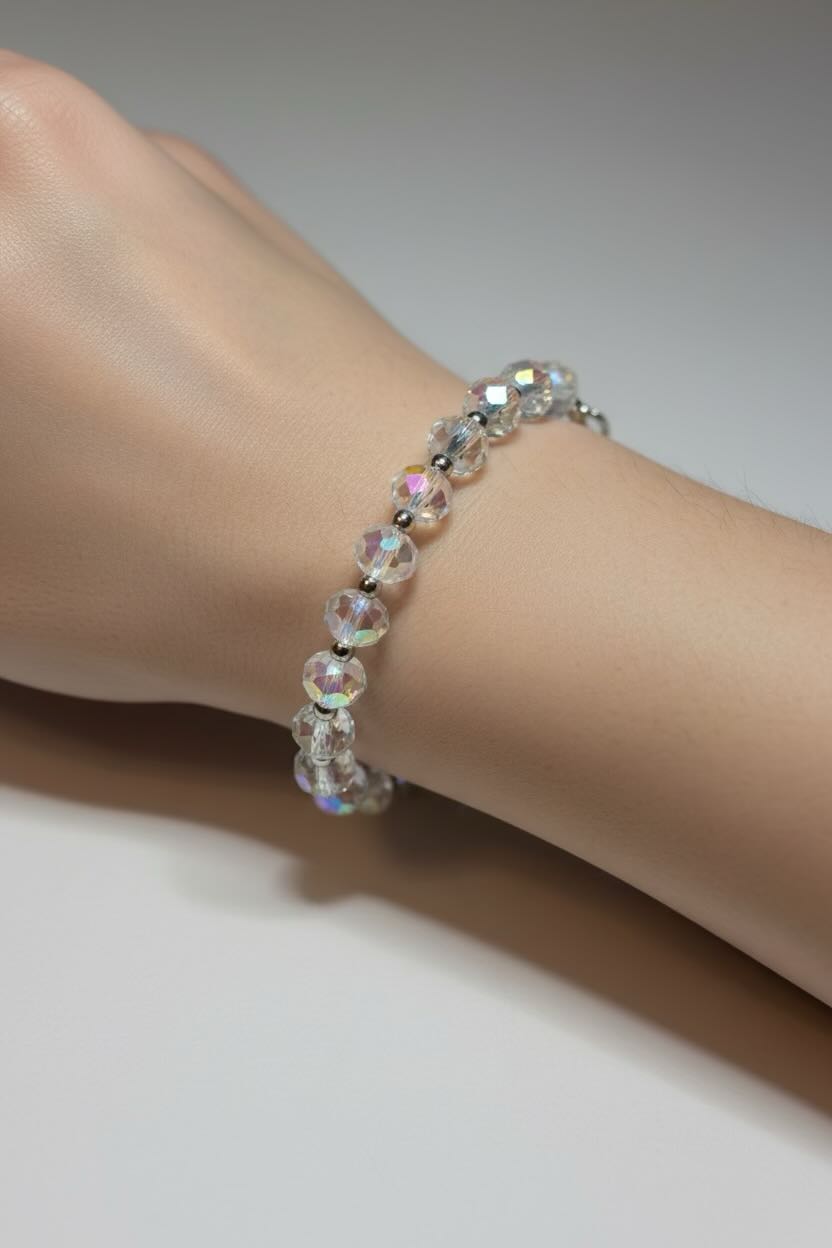 A close-up of a delicate beaded bracelet worn on a wrist. The bracelet features clear, faceted beads with an iridescent, rainbow-like sheen set in a silver-toned metal.