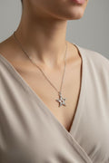 A close-up of a model wearing a delicate silver chain necklace with a star-shaped pendant, styled with a beige V-neck top against a neutral gray background.