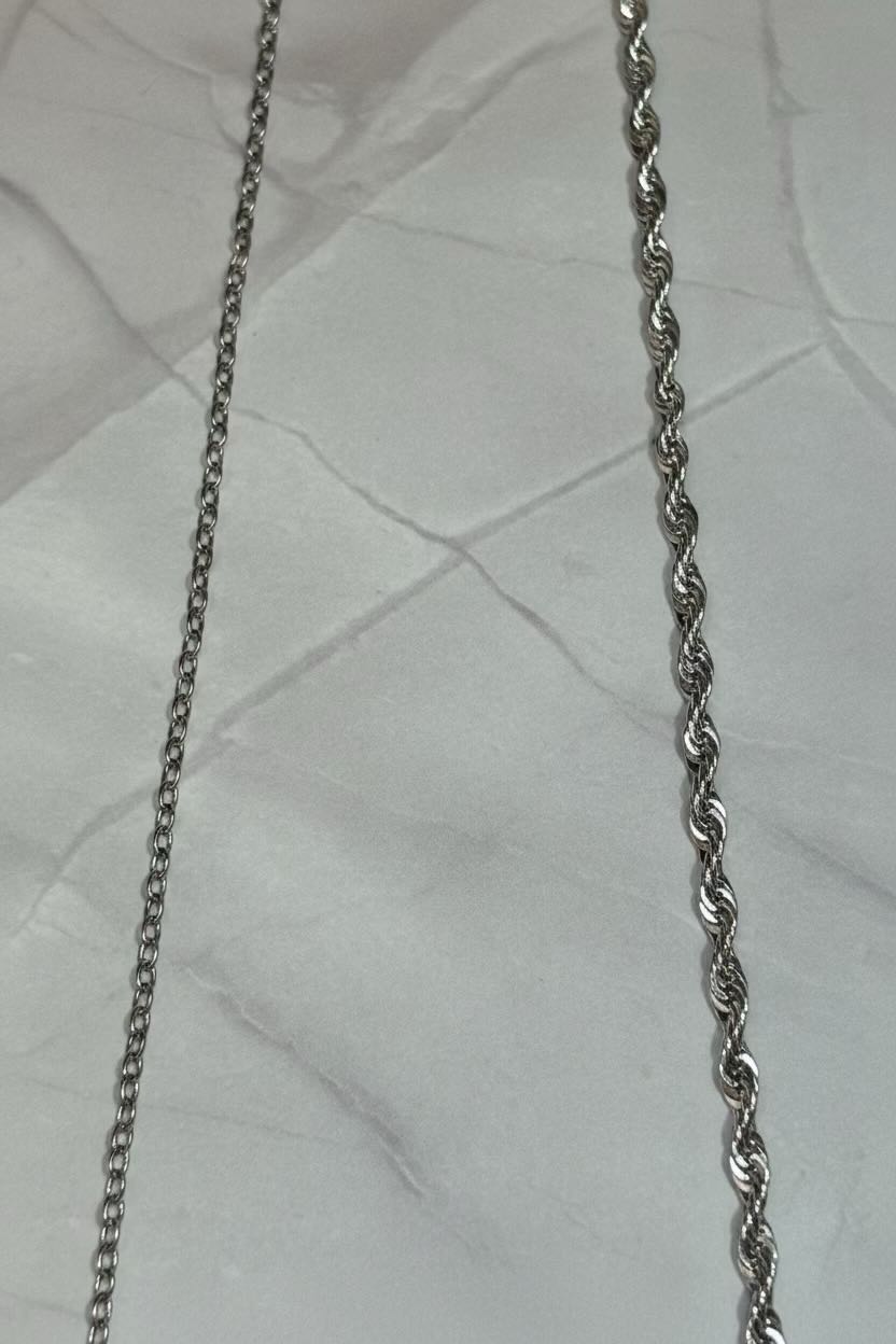 Two silver-toned metal chains laid flat on a white textured fabric background, one chain with a standard link pattern and the other with a twisted rope link pattern.