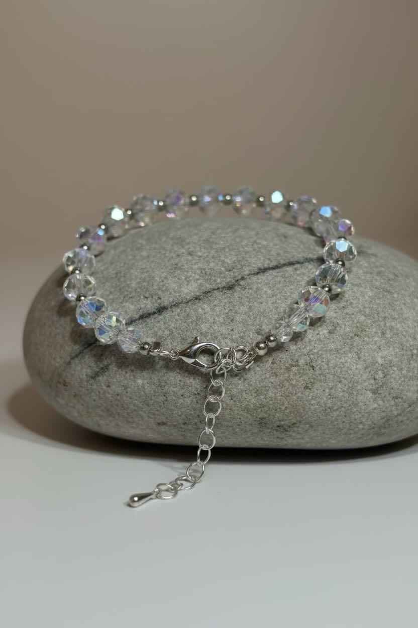 A silver-toned beaded bracelet with iridescent clear faceted beads, displayed on a smooth gray stone against a neutral beige background.
