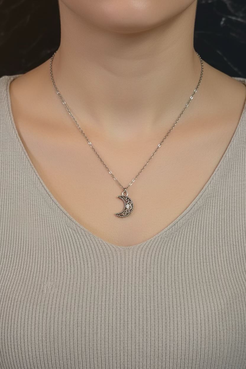 A silver crescent moon pendant necklace with small clear stones, worn on a model's neck over a light beige ribbed V-neck top.