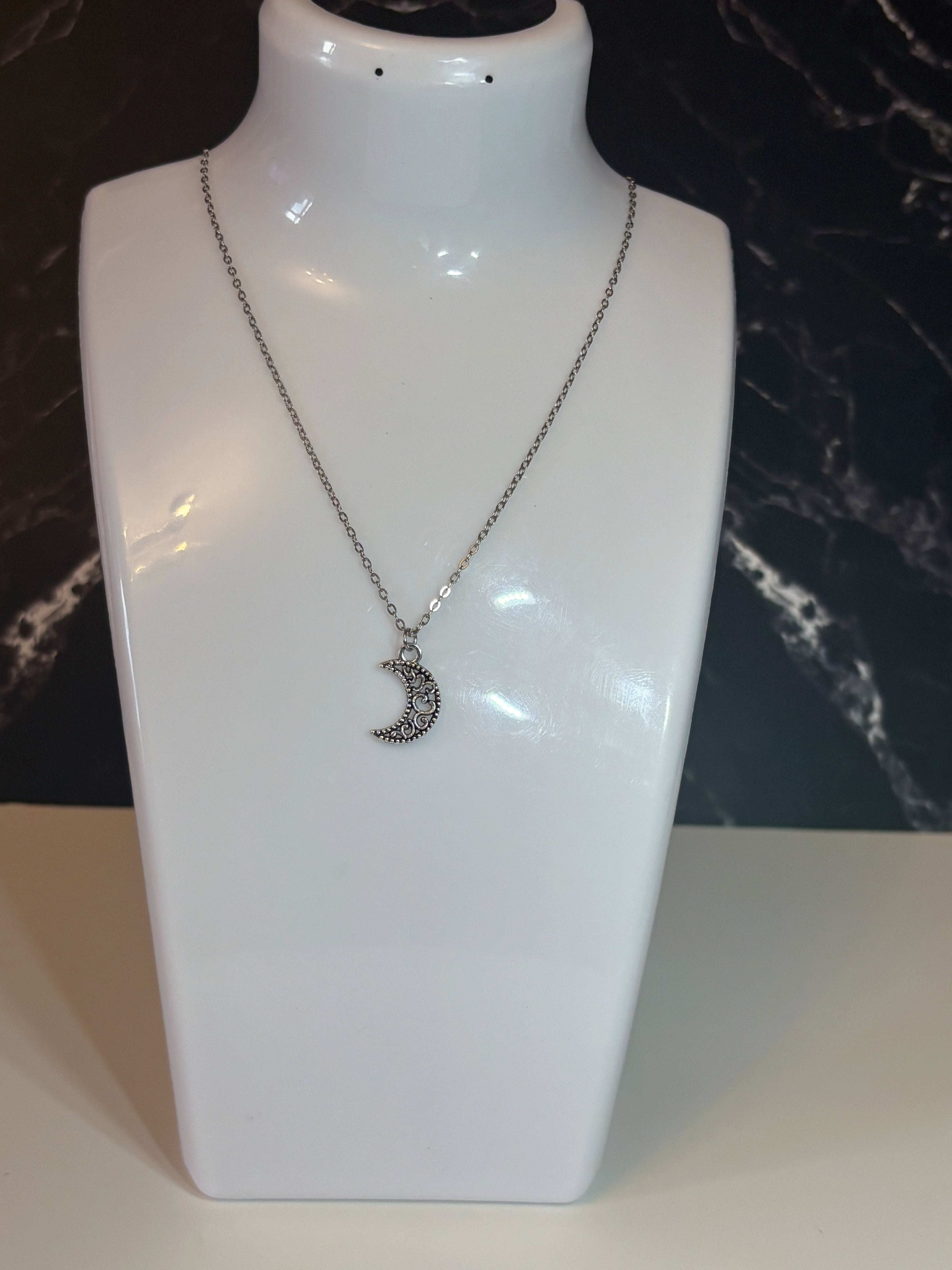 A silver crescent moon pendant necklace with small clear stones, displayed on a white jewelry bust against a dark marble-patterned background.