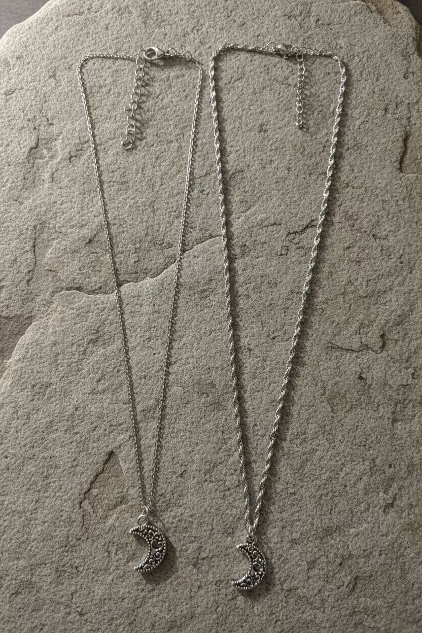 Two silver crescent moon pendant necklaces with delicate chains, displayed on a textured gray stone surface.