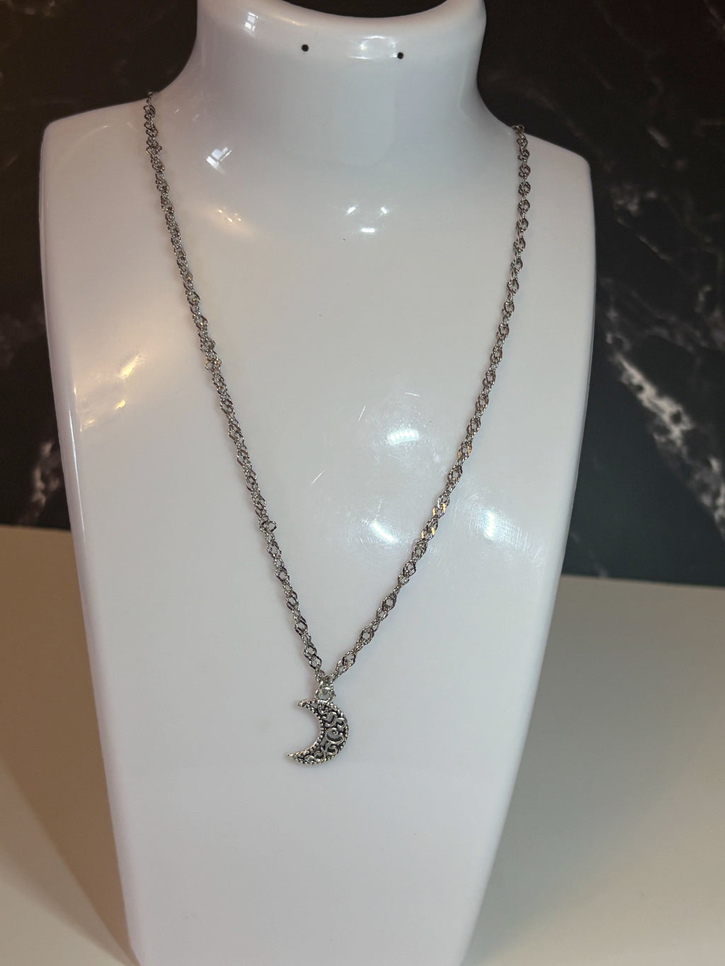 A close-up of a silver-toned chain necklace with a crescent moon pendant on a stand.