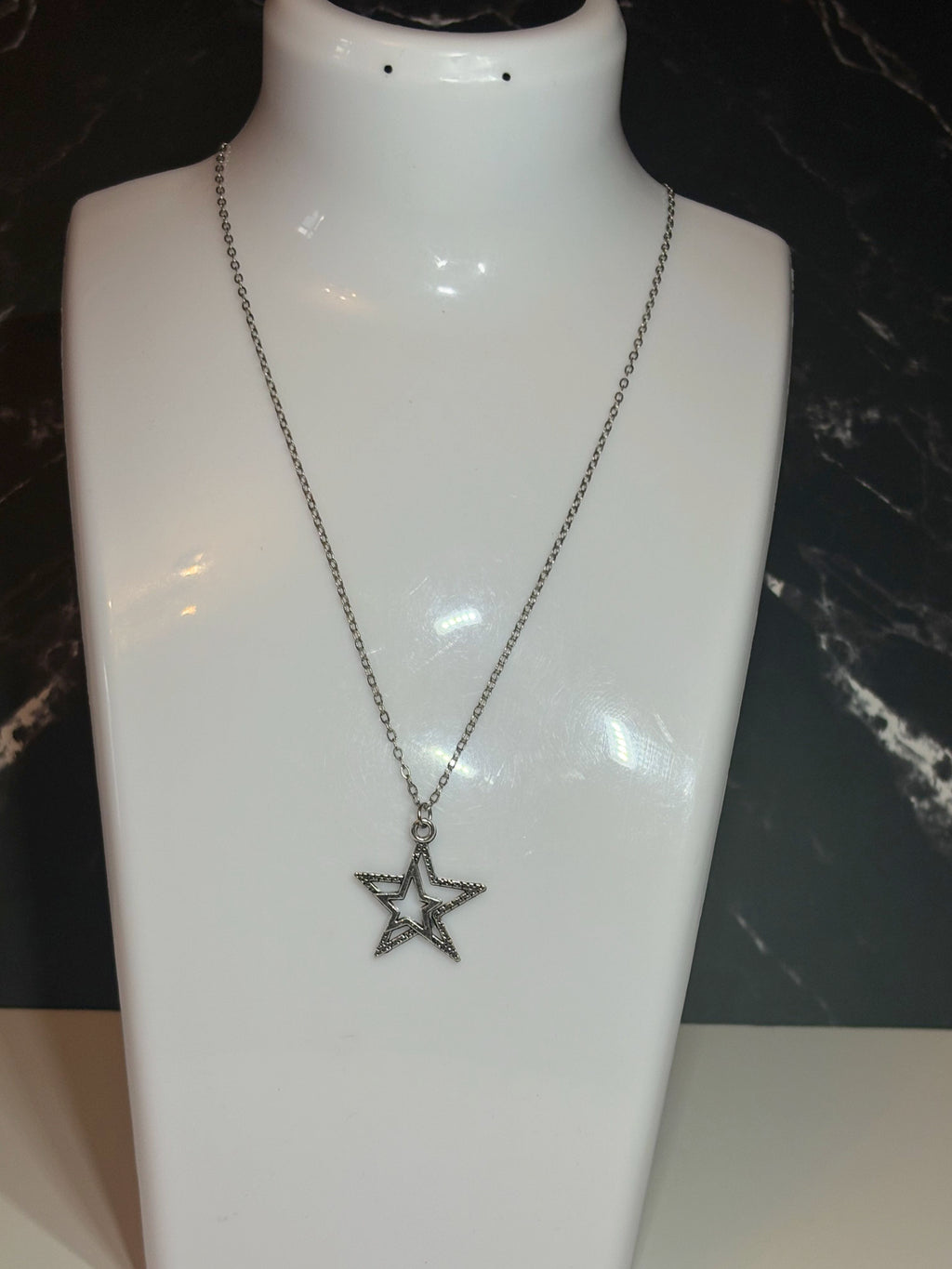A silver-toned star pendant necklace displayed on a glossy white jewelry bust against a dark marbled background.