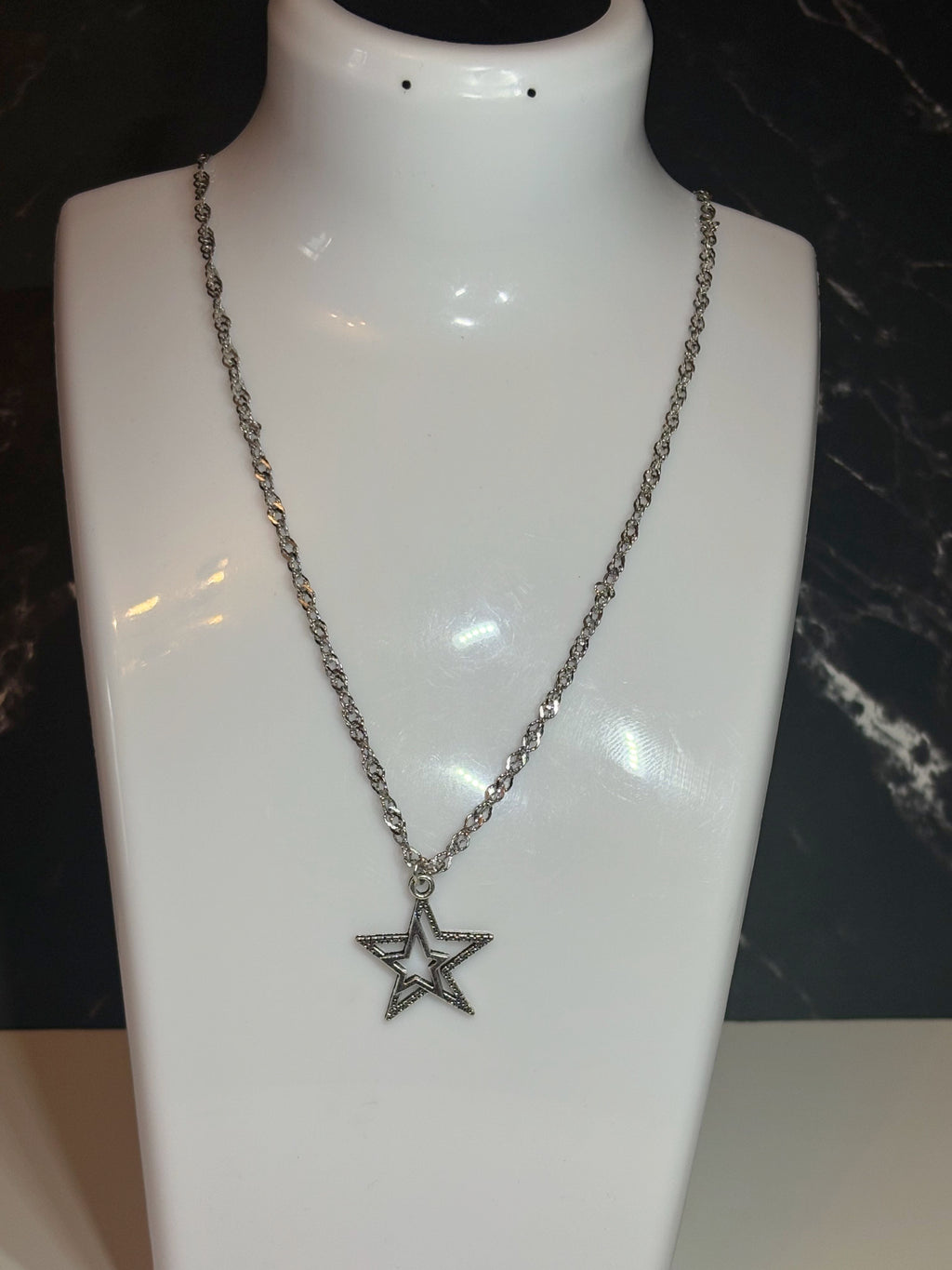 A silver-toned chain necklace with a star-shaped pendant, displayed on a glossy white mannequin bust against a dark background.