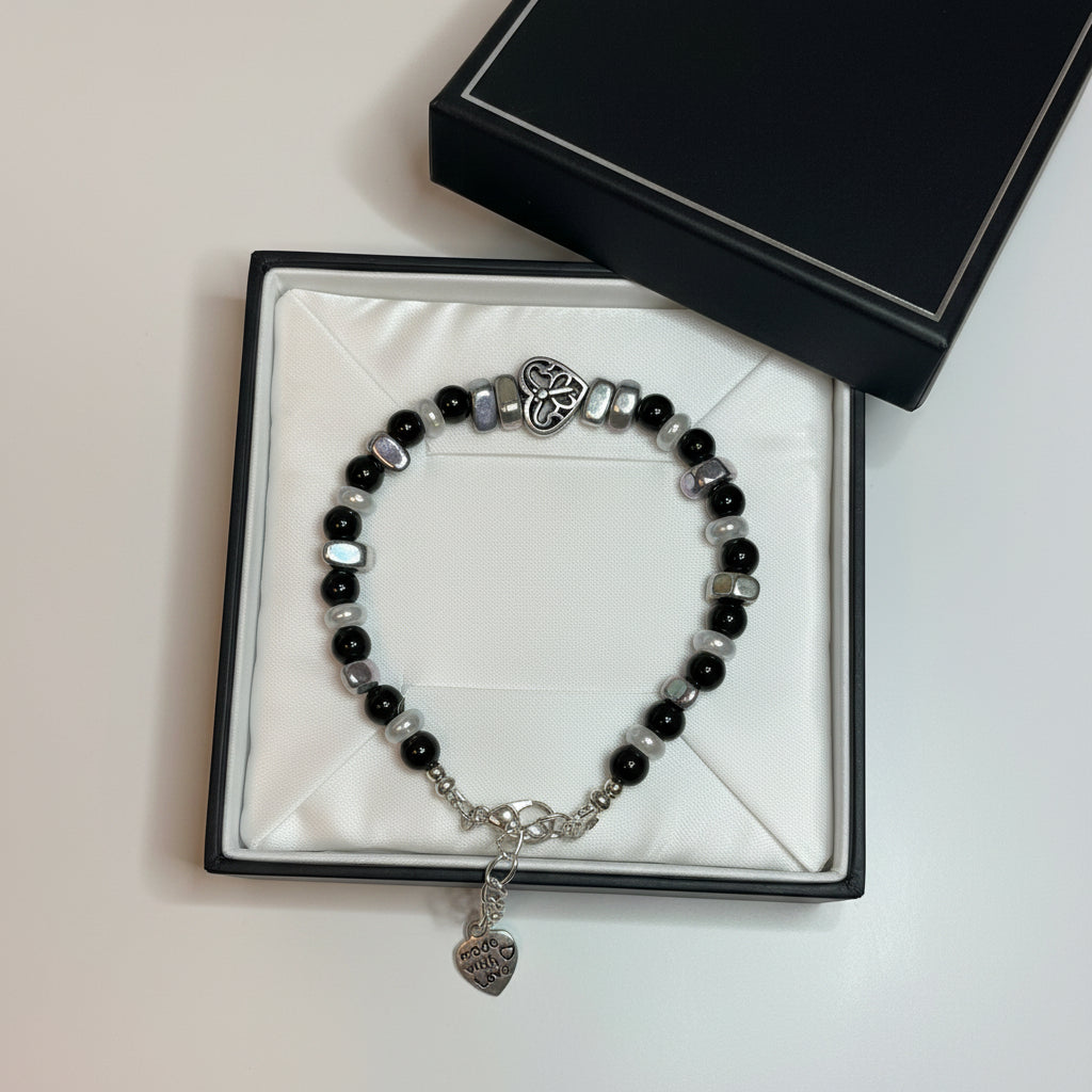 A beaded bracelet with alternating black and silver-gray glass stones, featuring a silver heart-shaped charm and a small dangling heart charm in a gift box.