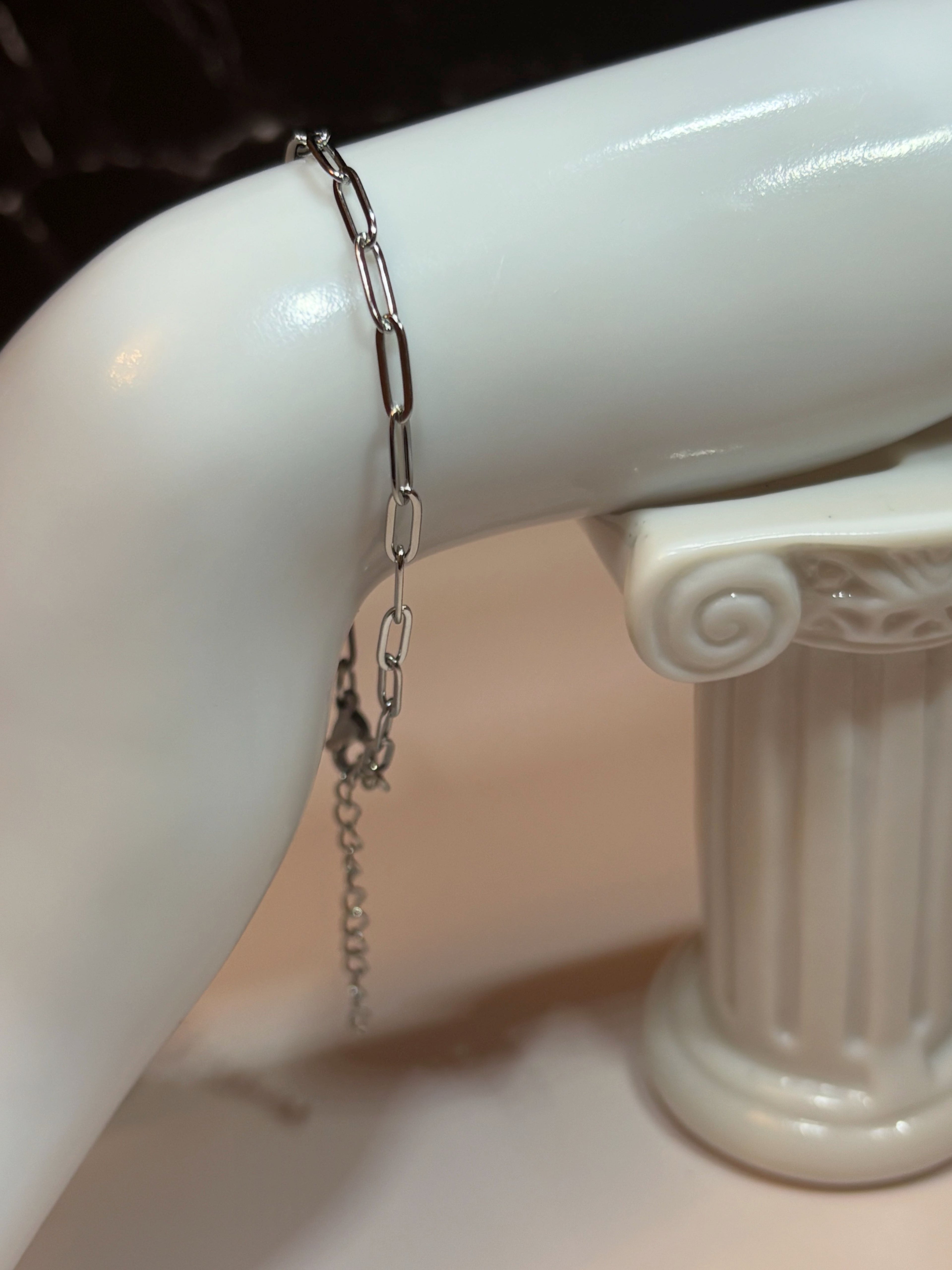 A silver paperclip chain bracelet displayed on a glossy white mannequin arm, with a matching clasp hanging down.