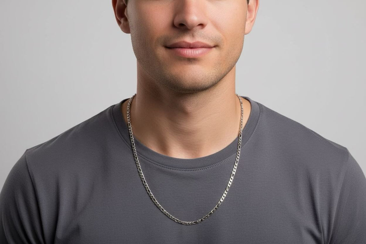 A close-up of a man wearing a silver chain necklace over a dark gray t-shirt, set against a plain light gray background.