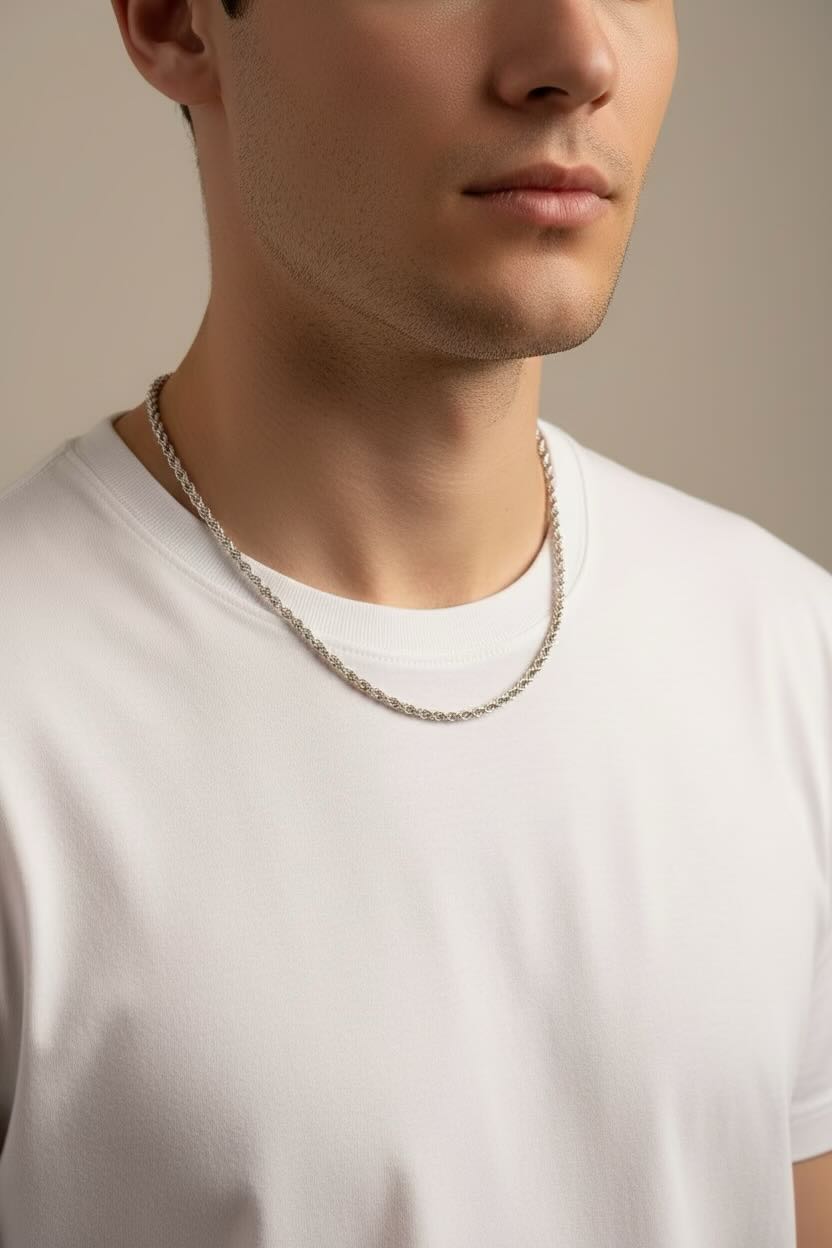 A close-up portrait of a man wearing a thin, polished silver chain necklace against a light gray background.