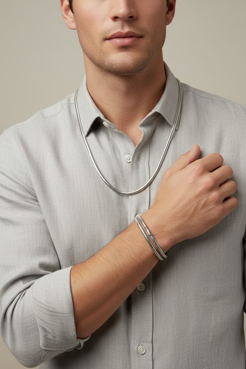A close-up of a man wearing a slim silver chain necklace and a matching silver bangle bracelet, paired with a light gray linen button-down shirt.