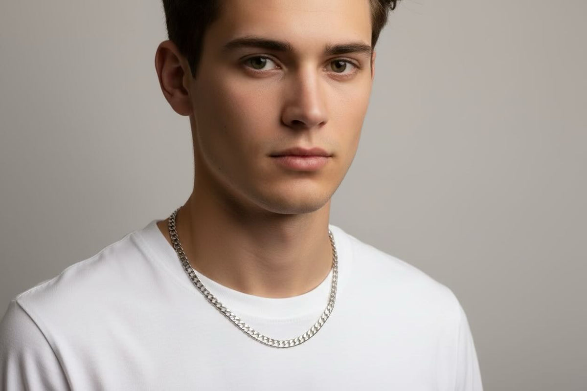 A close-up portrait of a young man wearing a silver chain necklace and a white t-shirt against a neutral gray background.