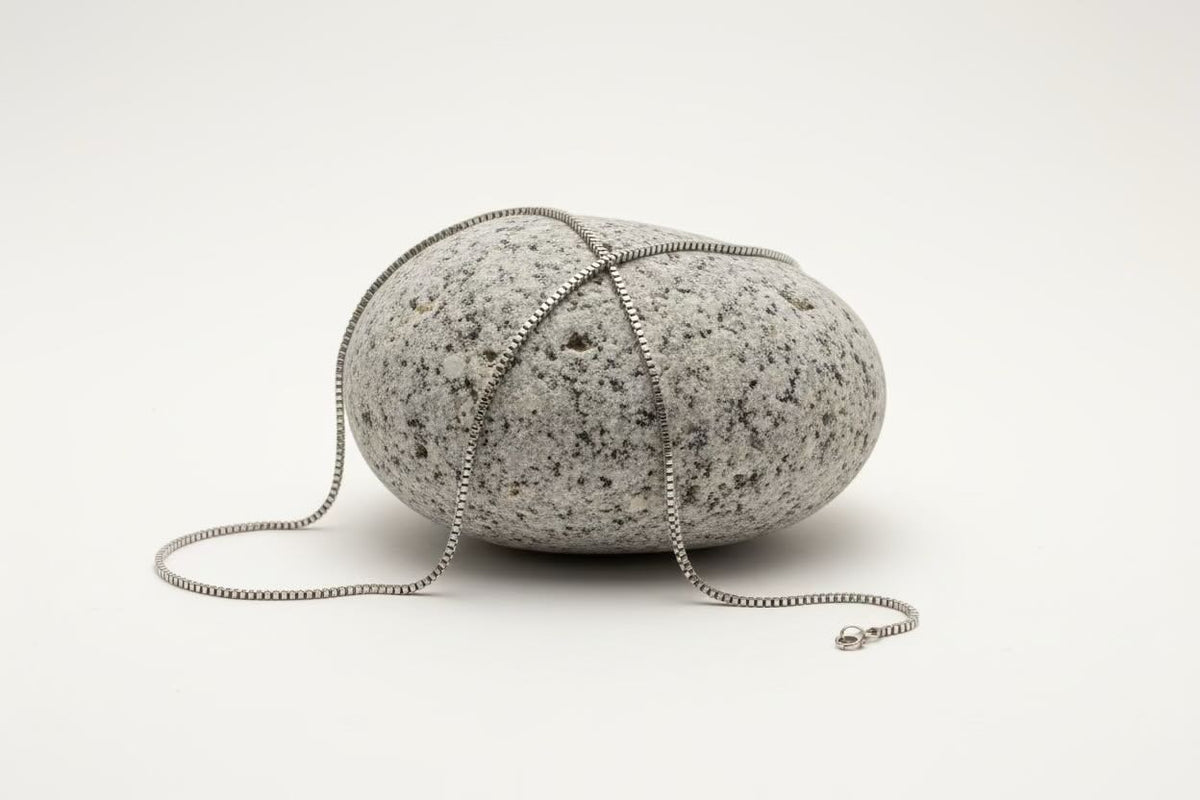 A silver-toned metal chain necklace draped over a smooth, rounded gray speckled stone, photographed on a clean white studio background.