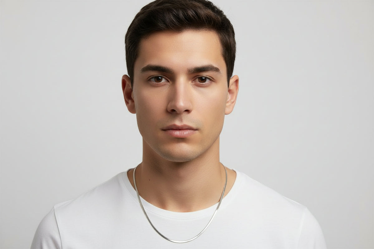 A close-up portrait of a man wearing a thin, polished silver chain necklace against a light gray background.