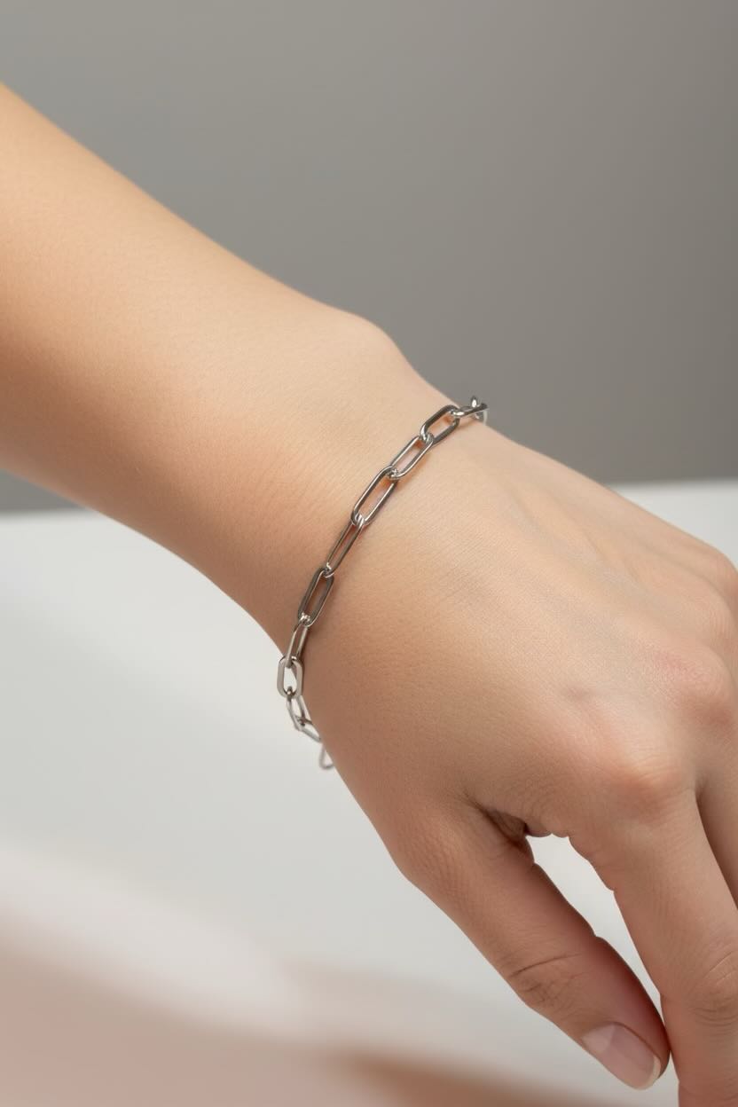 A close-up of a person's wrist wearing a delicate silver chain paperclip bracelet against a soft gray and white background.
