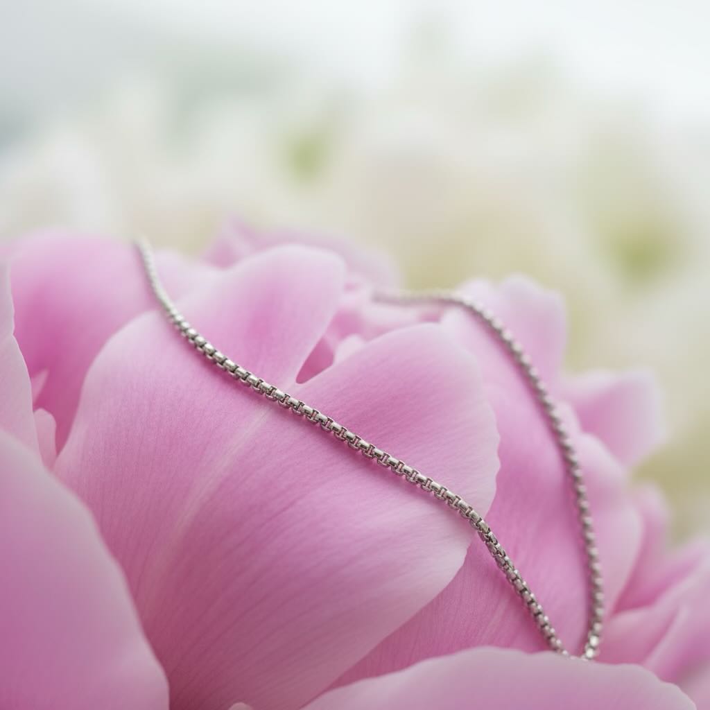 A delicate silver chain necklace draped across soft pink flower petals with a blurred white background.
