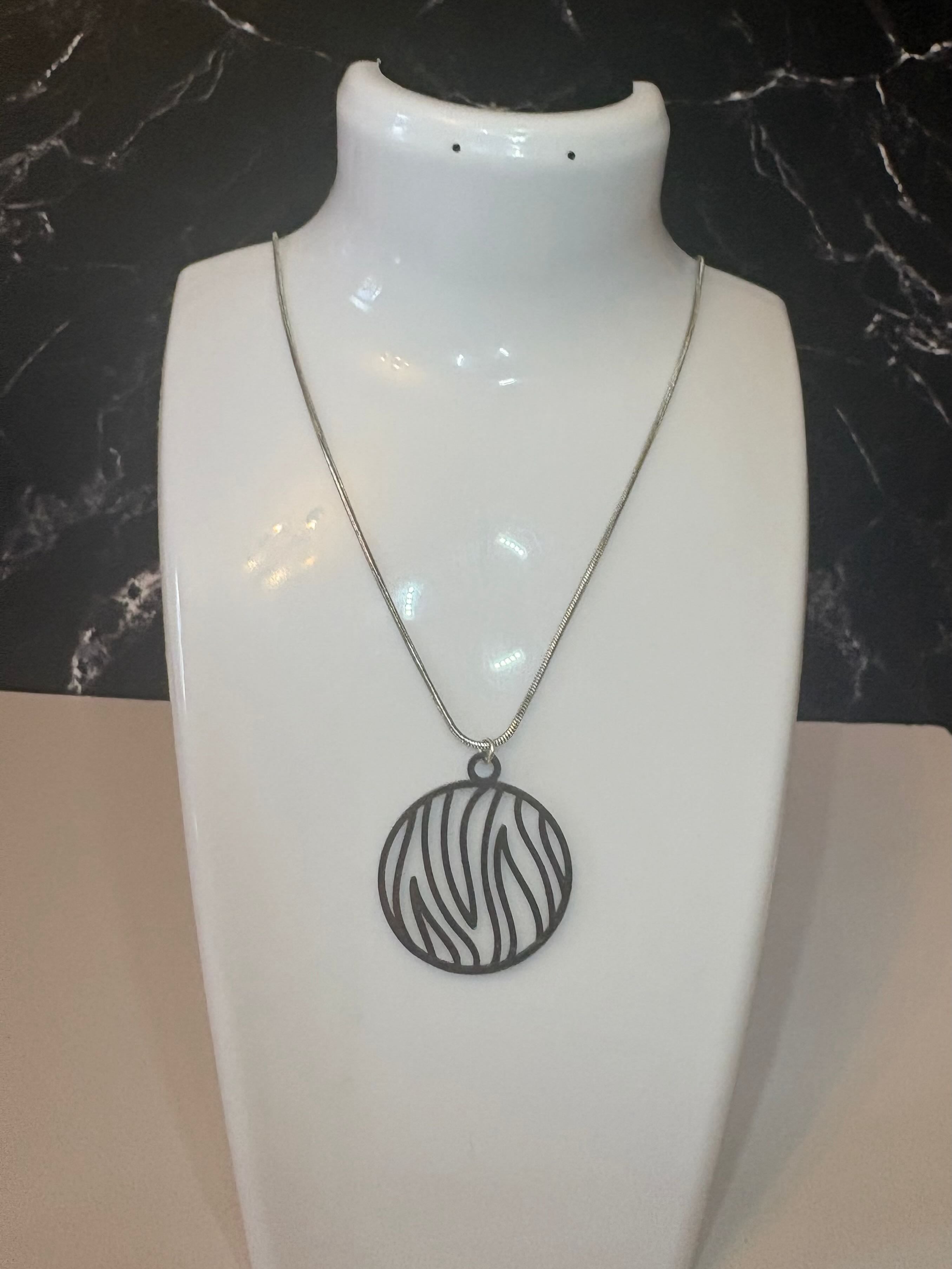 A silver-toned necklace with a circular pendant featuring a flame line pattern, displayed on a white jewelry bust against a dark marble-patterned background.