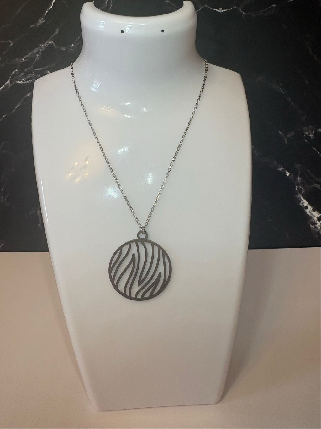 A silver-toned circular pendant necklace with a flame pattern, displayed on a white jewelry bust against a black marble background.