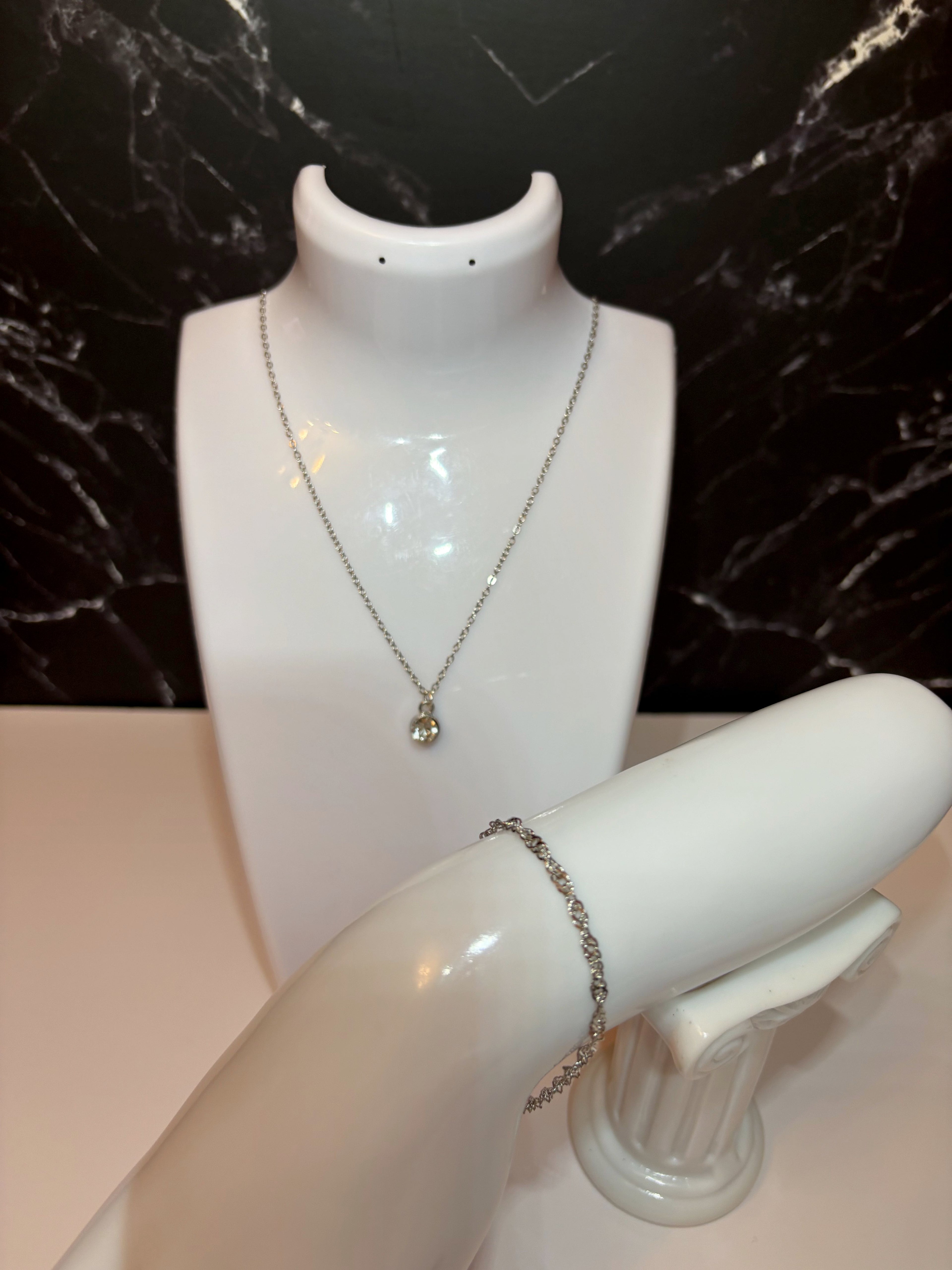 A silver chain necklace with a small diamond birthstone pendant displayed on a white jewelry bust, alongside a silver water wave chain bracelet on a white arm stand, set against a black marble background.