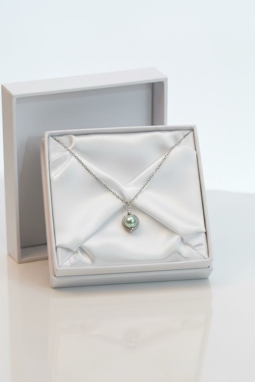 A silver necklace with a small round aqua blue pearl pendant, presented in an open white jewelry box with satin lining, with the box lid standing behind it on a white surface.