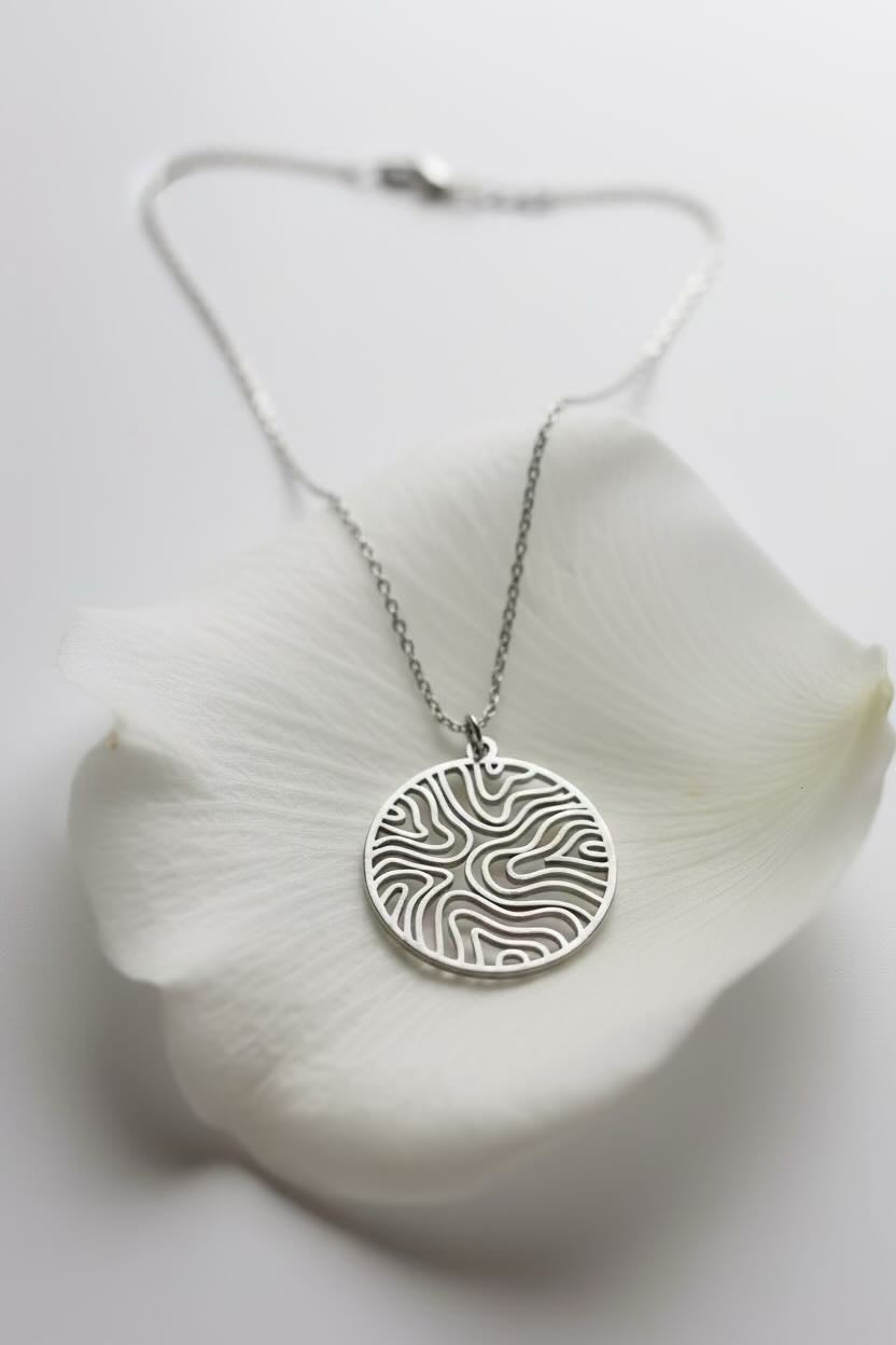 A close-up of a silver pendant necklace with a circular, wave pattern circle patterned pendant on a delicate silver chain, on a large white petal.