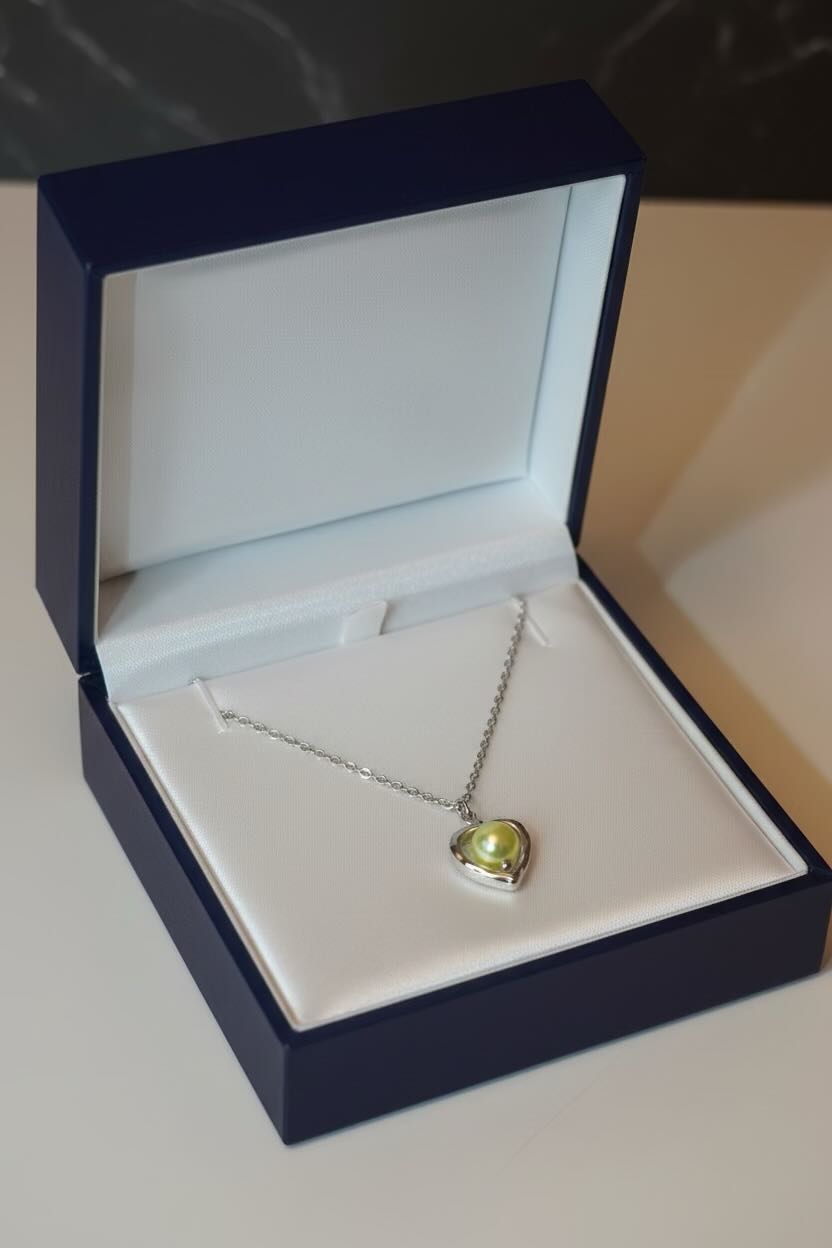 A silver heart pendant necklace with a champagne yellow pearl, displayed inside an open dark blue jewelry box with a white velvet insert.