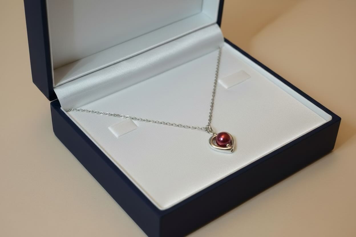 A silver heart pendant necklace with a cherry red pearl encased in a heart, displayed inside an open navy blue jewelry box with a white insert.