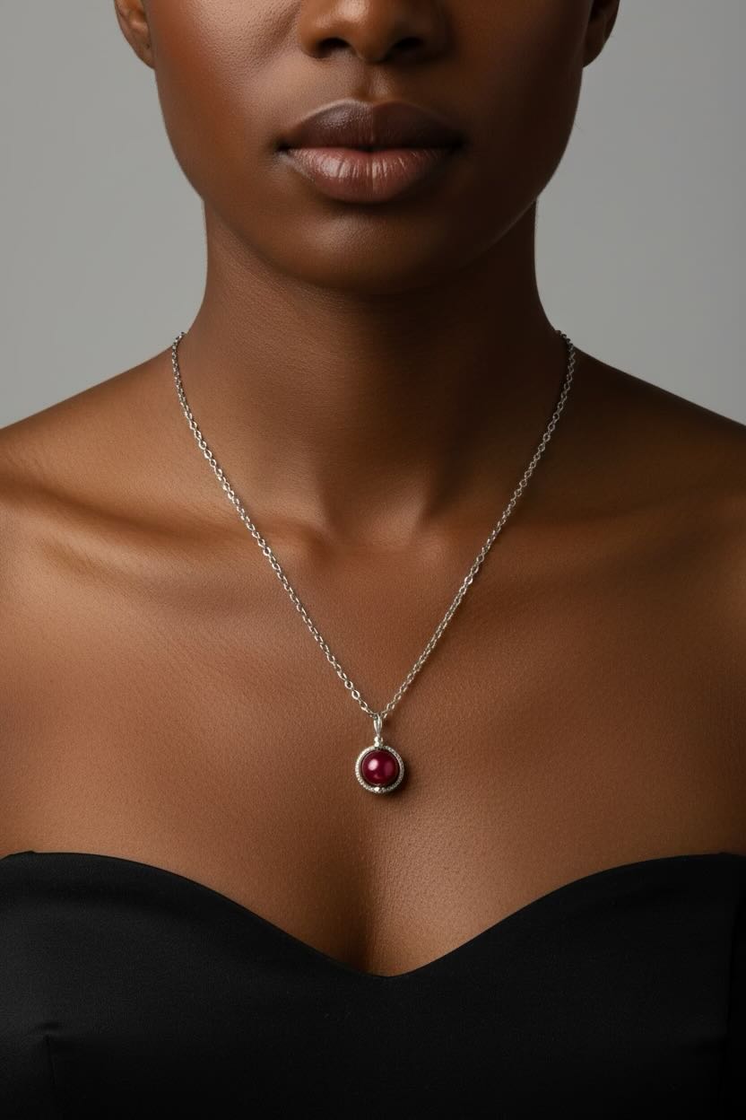 A close-up of a model wearing a delicate silver chain necklace with a round pendant featuring a cherry red pearl surrounded by a silver bezel.