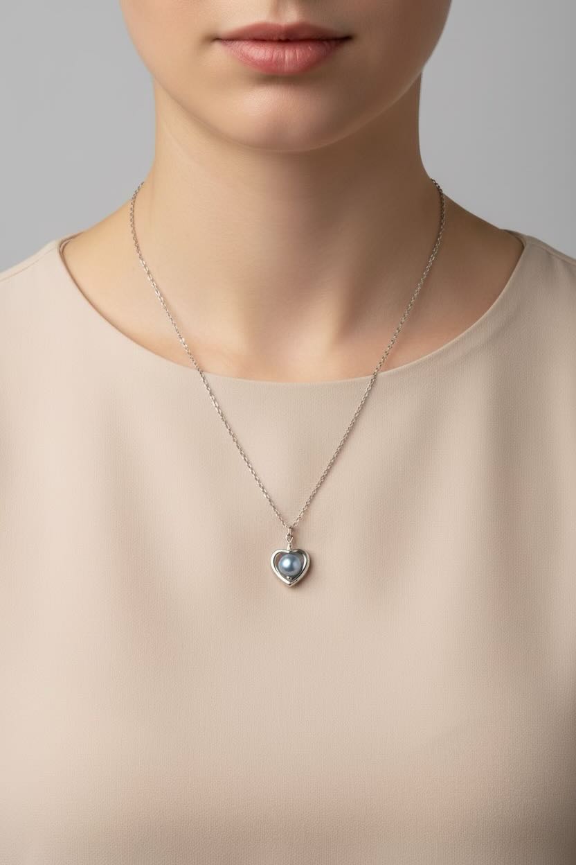 A close-up of a model wearing a delicate silver heart pendant necklace with a dark baby blue pearl styled with a beige top against a neutral gray background.