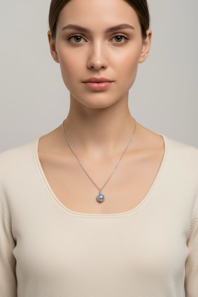 A woman wearing a delicate silver chain necklace with a round, dark baby blue pearl pendant, photographed against a neutral light gray background.