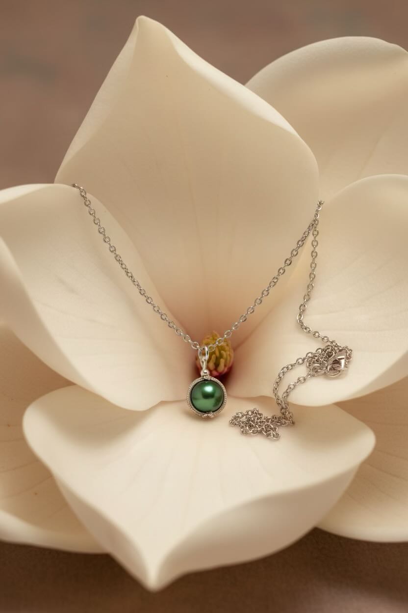 A delicate silver necklace with a dark green pearl pendant, displayed on the center petal of a large cream magnolia flower.