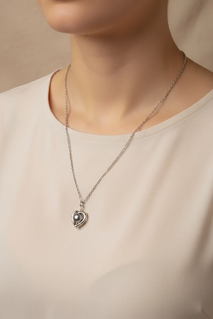 A close-up of a model wearing a delicate silver heart pendant necklace with a dark grey pearl in centre, styled with a simple cream-colored top.