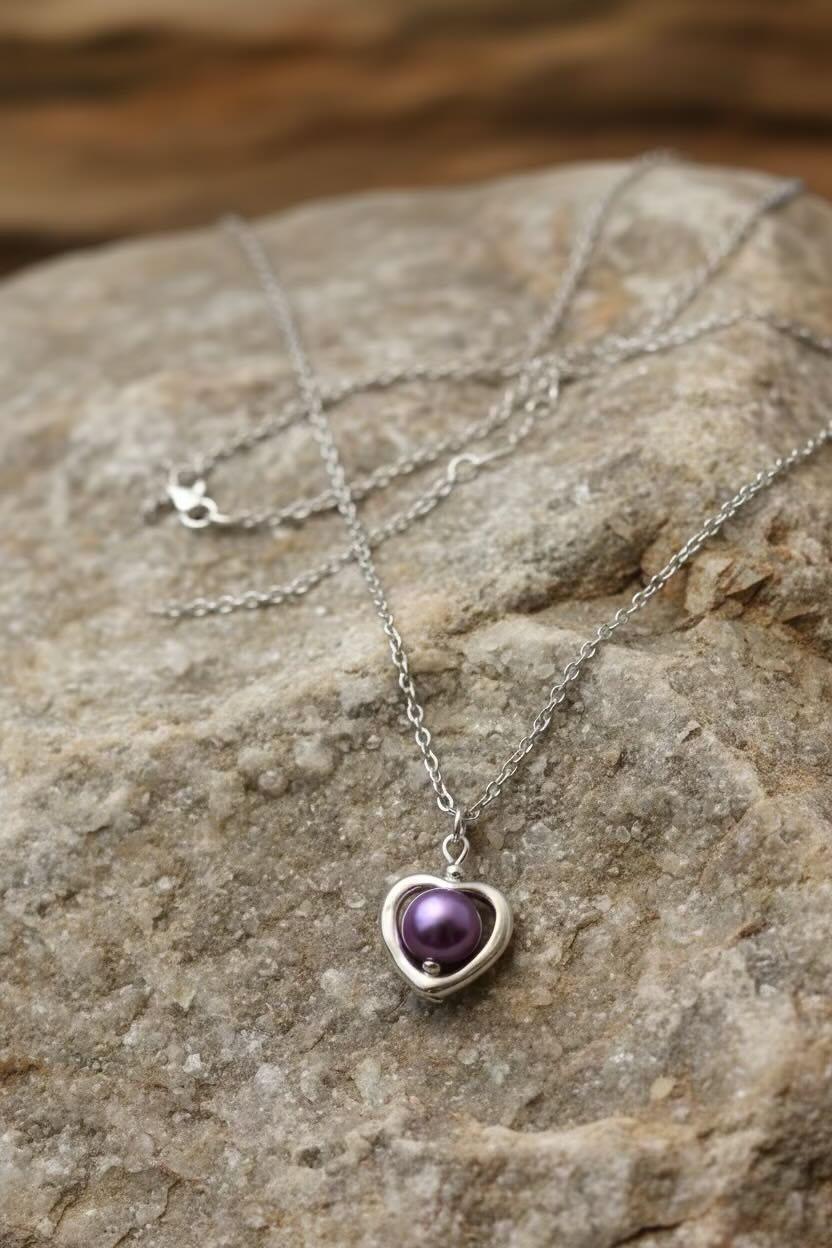 A silver heart pendant necklace with a dark orchid purple pearl in heart-shaped pendant, displayed on a textured gray rock.