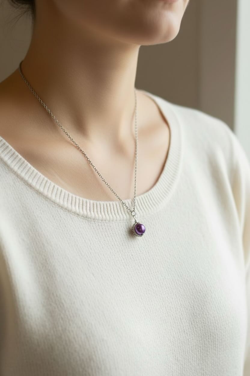A close-up of a person wearing a delicate silver chain necklace with a small round dark orchid purple pearl pendant, paired with a cream-colored knit top.
